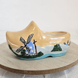 May include: A ceramic Dutch clog planter, painted with a windmill scene. The clog is tan with a blue and white windmill, green trees, and a blue base. It sits on a round wooden base.