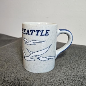 May include: A white ceramic mug with a speckled finish, featuring the word "SEATTLE" in navy blue and a design of flying seagulls. The handle and rim are also navy blue.