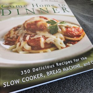 May include: A cookbook titled "Come Home to Dinner" features a photo of a plate of food covered in melted cheese. The book cover also includes the text "350 Delicious Recipes for the Slow Cooker, Bread Machine, and Oven."
