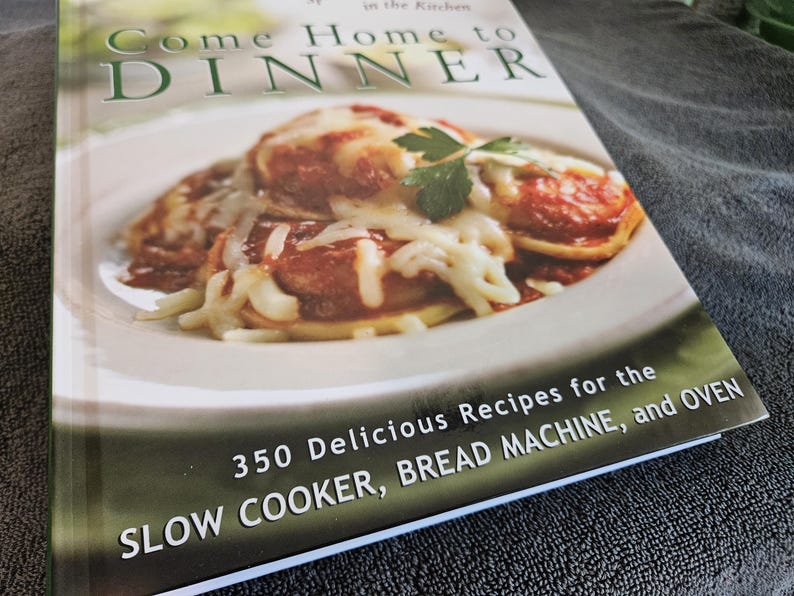 May include: A cookbook titled "Come Home to Dinner" features a photo of a plate of food covered in melted cheese. The book cover also includes the text "350 Delicious Recipes for the Slow Cooker, Bread Machine, and Oven."