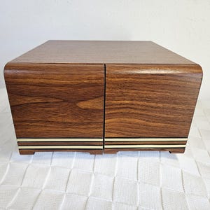 May include: A wooden box with a dark brown finish and a curved top. The box has two doors and a decorative cream and gold stripe along the bottom. The box is sitting on a white, textured surface.