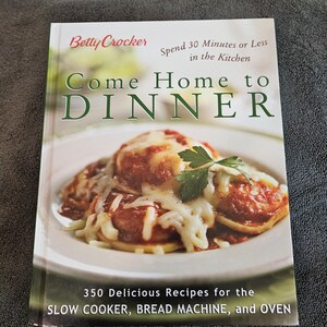 May include: A cookbook titled "Come Home to Dinner" by Betty Crocker. The cover features a photo of a plate of food covered in sauce and cheese. The book promises 350 recipes for slow cookers, bread machines, and ovens.