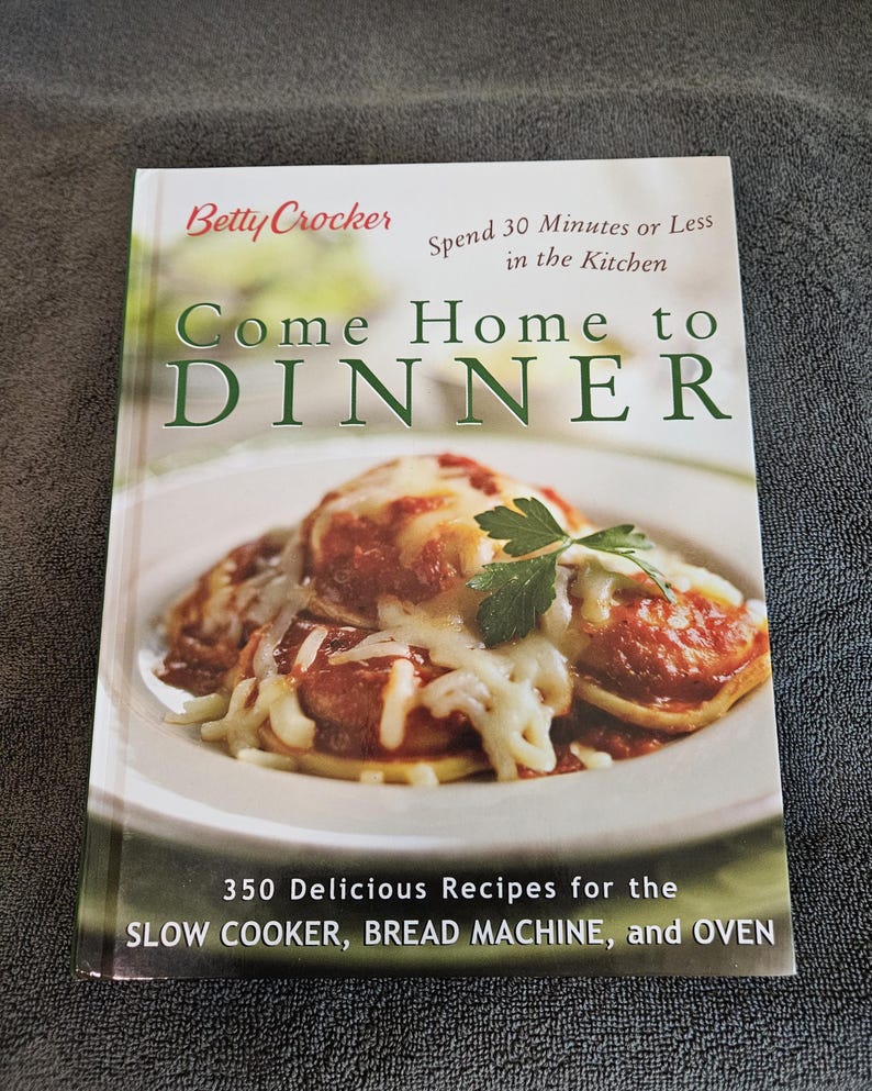May include: A cookbook titled "Come Home to Dinner" by Betty Crocker. The cover features a photo of a plate of food covered in sauce and cheese. The book promises 350 recipes for slow cookers, bread machines, and ovens.