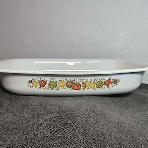 May include: White rectangular baking dish with a colorful fruit and vegetable design around the rim. The dish has a slightly curved shape with handles on each end. The design includes tomatoes, lemons, and other produce.