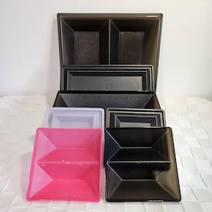 May include: A collection of plastic storage containers in various colors and sizes. The containers include black, pink, white, and gray options. The containers are square and rectangular, with some having divided compartments.