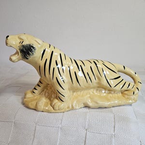 May include: A ceramic tiger figurine with black stripes on a cream background. The tiger is lying down with its mouth open and its head turned to the side.