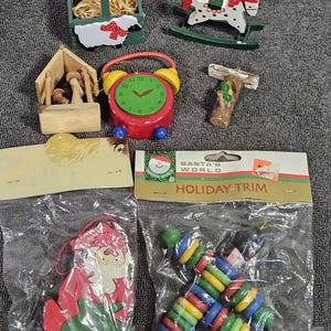 May include: A collection of Christmas ornaments, including a wooden rocking horse, a red and green alarm clock, and a wooden basket with a sheep design. Other ornaments include a wooden Santa Claus and a colorful beaded garland. "SANTA'S WORLD HOLIDAY TRIM" is printed on the packaging.