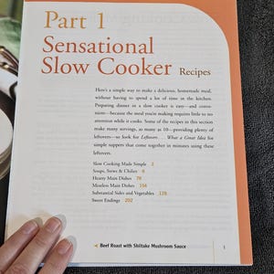 May include: Open cookbook page titled "Sensational Slow Cooker Recipes." The page has orange and white text. The book's table of contents includes sections on soups, stews, hearty main dishes, meatless dishes, sides, and desserts. A hand is visible.