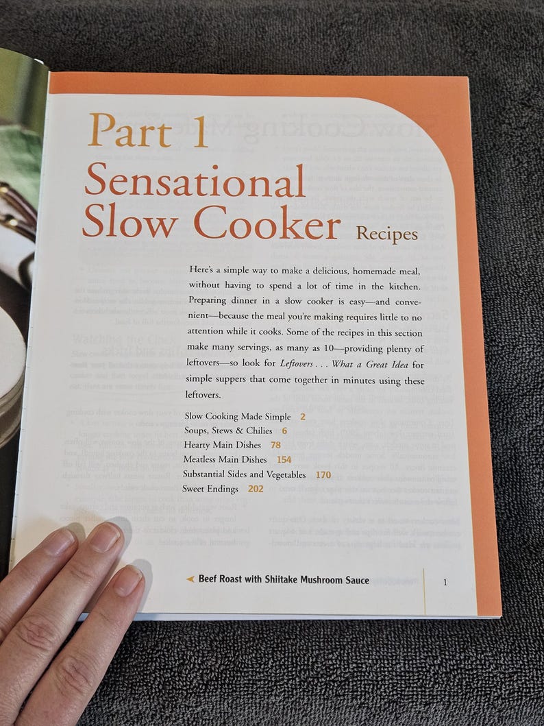 May include: Open cookbook page titled "Sensational Slow Cooker Recipes." The page has orange and white text. The book's table of contents includes sections on soups, stews, hearty main dishes, meatless dishes, sides, and desserts. A hand is visible.