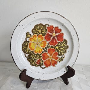 Pode incluir: Um prato de cerâmica branco com um desenho floral com flores laranja, amarelas e verdes. O prato tem um esmalte salpicado e uma borda marrom.