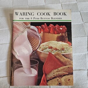 May include: Vintage cookbook titled "WARING COOK BOOK FOR THE 8 PUSH BUTTON BLENDER." The cover features a vibrant image of food, including a strawberry-topped cake, a bowl of food, and fried items. A pink liquid is being poured into a glass.
