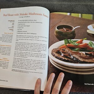 May include: A cookbook page open to a recipe for "Beef Roast with Shiitake Mushroom Sauce." A plate of sliced roast beef with mushroom sauce, carrots, and snap peas sits on a stack of plates. A small bowl of sauce and a spoon are visible.