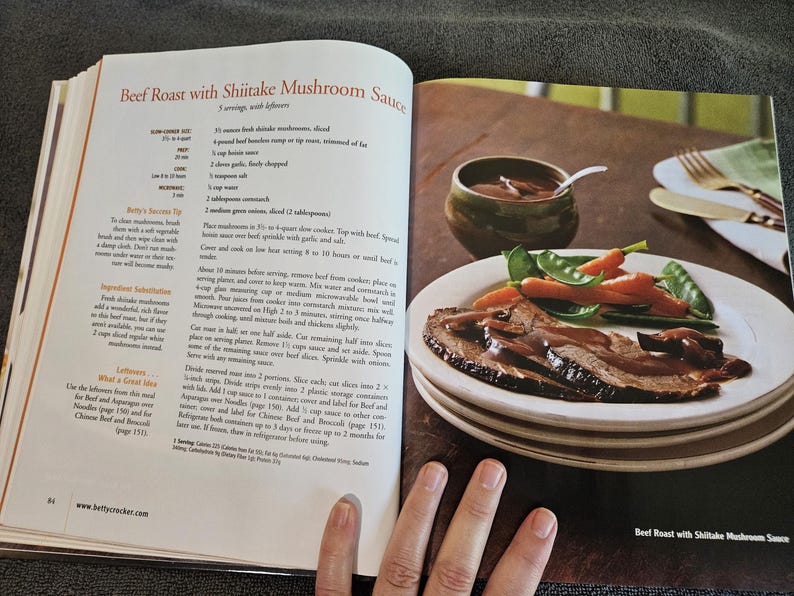 May include: A cookbook page open to a recipe for "Beef Roast with Shiitake Mushroom Sauce." A plate of sliced roast beef with mushroom sauce, carrots, and snap peas sits on a stack of plates. A small bowl of sauce and a spoon are visible.