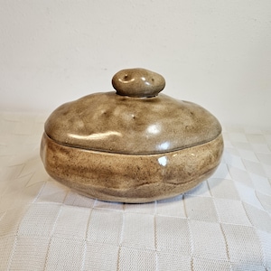 May include: A round, lumpy, potato-shaped ceramic lidded container with a mottled, earthy brown and tan glaze. The surface features an uneven, organic texture with small indentations and a small, rounded knob handle on the center of the lid.