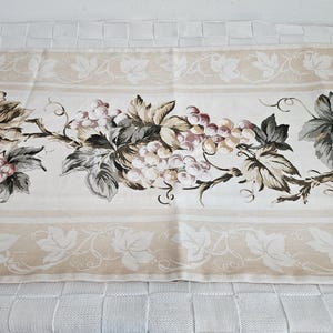 May include: A beige fabric table runner with a repeating pattern of white grape clusters and green leaves. The runner has a white border with a delicate vine design.