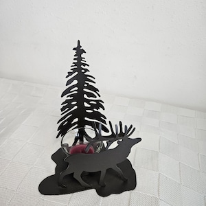 May include: Black metal candle holder featuring a silhouette of a pine tree and a deer. The candle holder has a glass cup to hold a candle. The candle holder is on a black base.