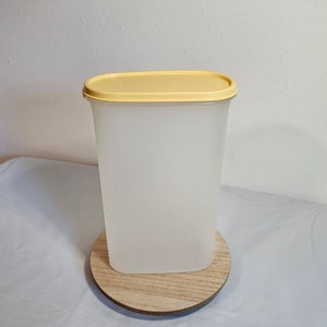 Könnte beinhalten: Ein rechteckiger Behälter aus klarem Kunststoff mit einem gelben Deckel. Der Behälter steht auf einem Holzsockel.