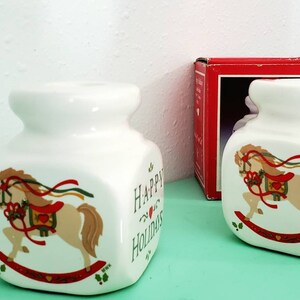 May include: Two white ceramic candle holders with a rocking horse design and the text "Happy Holidays" on the front. The candle holders are on a green surface.