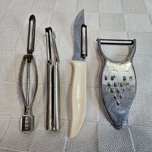 May include: A collection of kitchen tools including two metal peelers, a serrated knife with a white handle, and a metal grater. The tools are arranged on a white patterned surface.