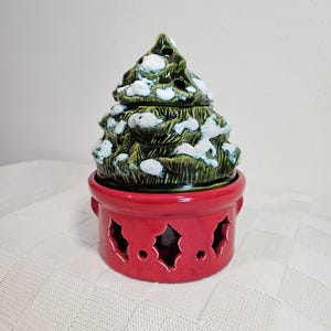 May include: A ceramic Christmas tree with a green, snow-covered design. The tree sits on a red base with holly-shaped cutouts. The base has small handles on either side.