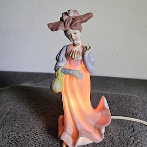 Può includere: Una lampada a forma di statuetta in porcellana raffigurante una persona con un cappello color malva a tesa larga, un corpetto color lavanda con dettagli dorati e una lunga gonna color pesca. La gonna si illumina dall'interno e pende un sacchetto.