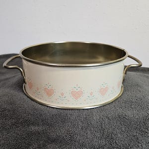 May include: A white enamelware bowl with a silver rim and handles. The bowl is decorated with a repeating pattern of pink hearts and green floral accents. The interior of the bowl is a gold color.
