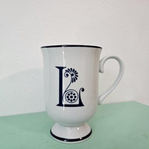 Pode incluir: Caneca de cerâmica branca com um design floral azul e branco e a letra "L" na frente. A caneca tem uma base de pedestal.