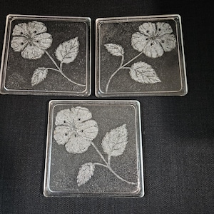May include: Three square glass coasters with an etched floral design. Each coaster has a flower and leaves. The coasters are clear and rest on a black surface.