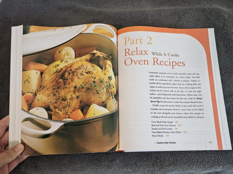 May include: Open cookbook page with a roasted chicken and vegetables in a pot. The text reads "Part 2 Relax While It Cooks Oven Recipes". The image is a close-up of a cooked chicken with potatoes and carrots.