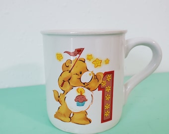 May include: White ceramic mug with a Care Bear holding a cupcake with a candle on top. The bear is wearing a red hat with a white flag. The mug has a large number "1" with yellow stars around it. The mug is on a light green surface.
