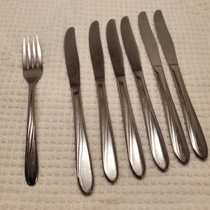 May include: A set of seven silver-toned flatware pieces, including six knives and one fork. The knives have a slightly curved handle and a pointed tip. The fork has four tines and a curved handle.