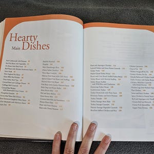 May include: Open cookbook pages with a list of recipes. The left page has the title "Hearty Main Dishes" with a list of recipes below. The right page has a list of recipes. The pages are white with orange accents.