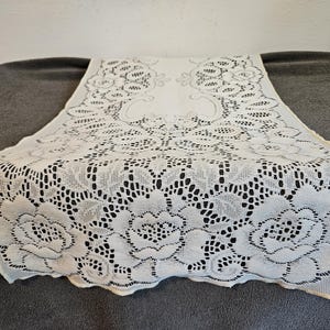 May include: A rectangular off-white lace table runner featuring an intricate floral pattern with detailed rose motifs and openwork mesh sections. The edges are scalloped, and the design is symmetrical, showcasing a repeating botanical lace weave.