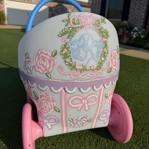 May include: A white toy baby carriage featuring a blue handle and pink wheels. The exterior is decorated with hand-painted pink roses, green leaves, and light pink bows. A central wreath design surrounds a blue monogrammed letter R on the front.