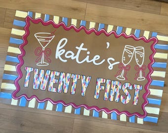 May include: A rectangular sign with a brown background, featuring the text "katie's TWENTY FIRST" in white and colorful letters. The sign includes illustrations of a martini glass and champagne flutes, all outlined in white with pink bows.