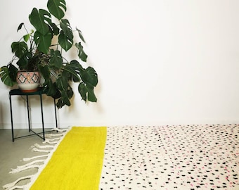 May include: A white and yellow rug with black and pink polka dots. The rug has a fringe border and is laid out on a light wood floor. A potted plant is in the background.