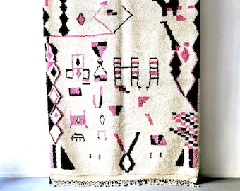 Könnte beinhalten: Ein weißer Teppich mit einem geometrischen Muster in Schwarz und Rosa. Der Teppich hat einen Fransenrand und hängt an einer Wand.