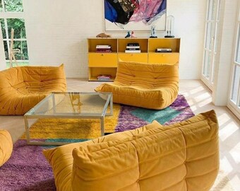 Könnte beinhalten: Ein modernes Wohnzimmer mit senfgelben modularen Sofas, einem Couchtisch mit Glasplatte und einem farbenfrohen geometrischen Teppich. Ein gelber Schrank mit Büchern und Dekoration befindet sich unter einem abstrakten Gemälde. Der Raum hat weiße Wände und große Fenster.