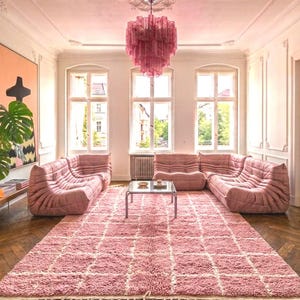Könnte beinhalten: Ein Wohnzimmer mit einem flauschigen, rosafarbenen Teppich mit Gittermuster und Fransen, drei niedrigen, gesteppten modularen Sofas in Rosa, einem Couchtisch mit Glasplatte und einem gestuften, spitzenartigen Kronleuchter in Rosa.