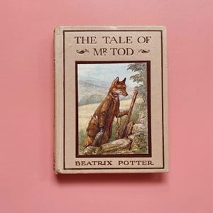 May include: A vintage book titled "The Tale of Mr. Tod" by Beatrix Potter. The cover features an illustration of a fox in a brown coat, holding a walking stick, set against a landscape background. The book has a beige cover and is set against a pink background.