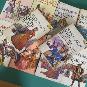 May include: A selection of vintage Jean Plaidy paperback books. The covers display illustrations of historical figures in period dress. Titles include "The Sixth Wife" and "Royal Road to Fotheringay". The books are arranged on a green surface.