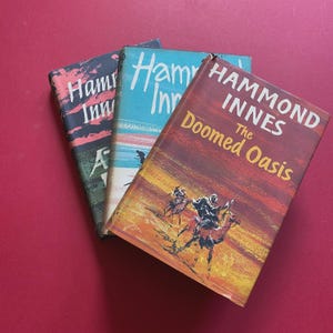 May include: Three vintage hardcover books with the author's name "Hammond Innes" visible on the spines. The book covers are in shades of red, blue, and yellow, with illustrations and text. The book titled "The Doomed Oasis" is visible. The books are on a red surface.