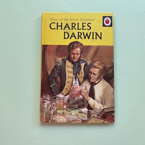 May include: A vintage book titled 'Lives of the Great Scientists' featuring Charles Darwin. The cover is yellow with black text and an illustration of two men examining a brain model. The background is a light teal colour.