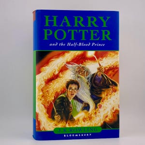 Puede incluir: Un libro de tapa dura titulado "Harry Potter y el Misterio del Príncipe" de J.K. Rowling. La portada presenta una ilustración de Harry Potter, un mago y llamas. El libro tiene una columna azul y un borde verde.