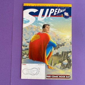 May include: Superman comic book cover featuring Superman in a red and yellow cape, blue suit, and the Superman logo. The cover has the text "Superman" in large white letters, with the DC Comics logo. The background shows a city and clouds.