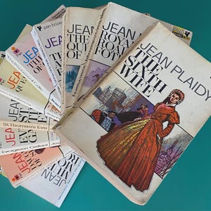 May include: A selection of vintage Jean Plaidy paperback books, arranged in a fan shape. The covers display illustrations and titles including "The Sixth Wife" and "The Road to Fotheringhay". The books are from the Pan Books series.