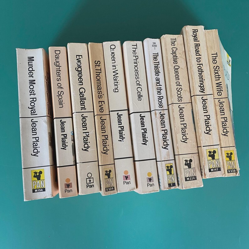 May include: A selection of vintage Jean Plaidy paperbacks, including titles such as "Murder Most Royal" and "The Sixth Wife." The books have off-white spines with black text and are displayed on a teal surface.