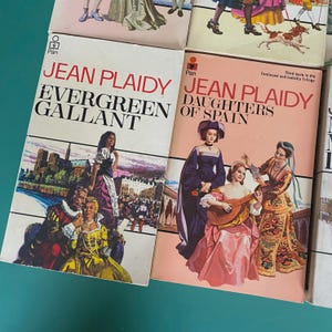 May include: A collection of vintage Jean Plaidy paperback books. The covers display illustrations of historical scenes and figures. Titles include "Evergreen Gallant" and "Daughters of Spain". The books have a retro design with a pink and white colour scheme.