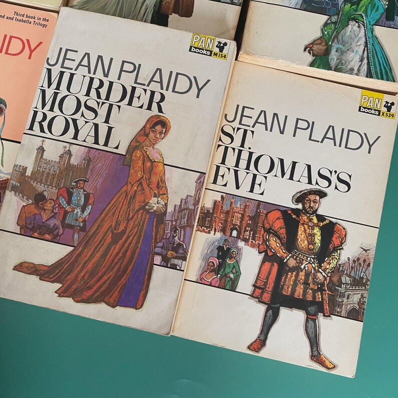 May include: A collection of vintage Jean Plaidy paperback books. The covers showcase illustrations of historical figures and scenes. Titles include "Murder Most Royal" and "St. Thomas's Eve," with the publisher's logo "PAN books" visible.