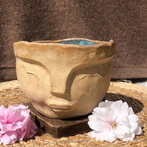 May include: A ceramic planter shaped like a face with closed eyes and a small wooden base. The planter is beige with a blue glaze on the inside. The planter is sitting on a woven mat with pink flowers.